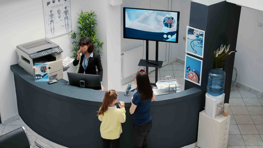 Receptionist working in hospital waiting area and offering support to patients at checkup visit, mother with child having medical appointment with physician. Reception desk at healthcare clinic.